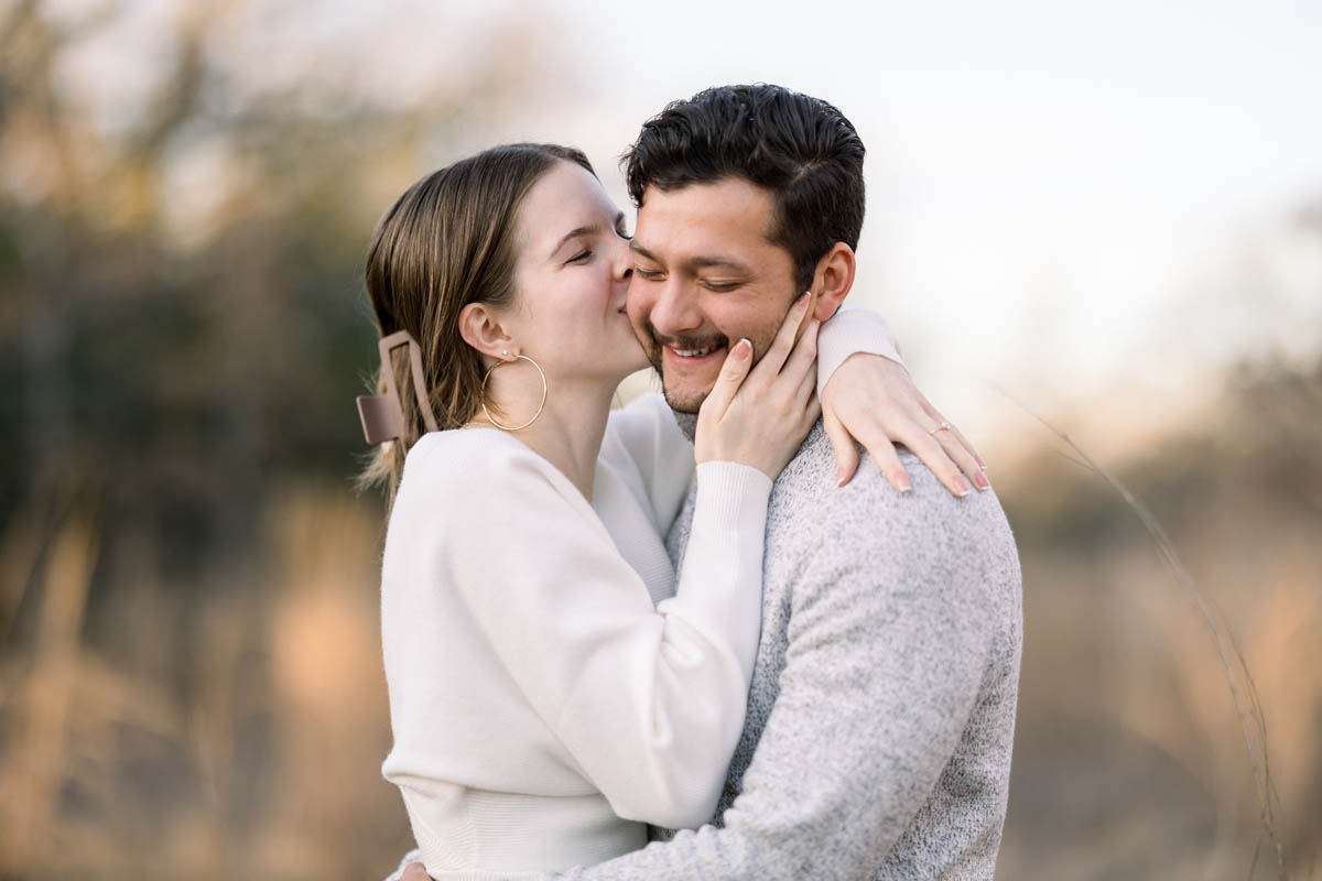 Woman in white kissing a man on the cheek while laughing in a brightly lit field